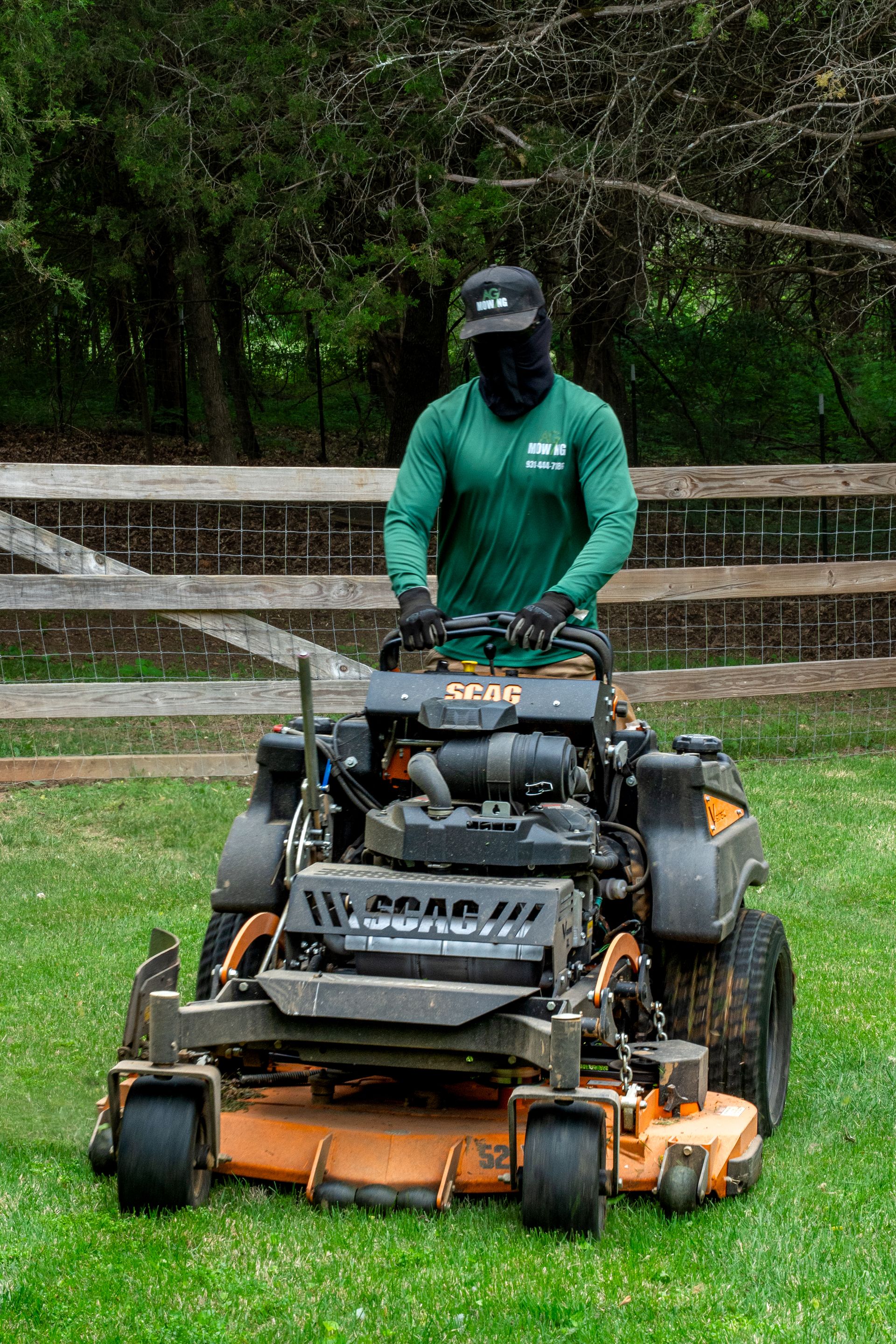 Person mowing a grassy field on a riding lawn mower, wearing a green shirt and black face covering.