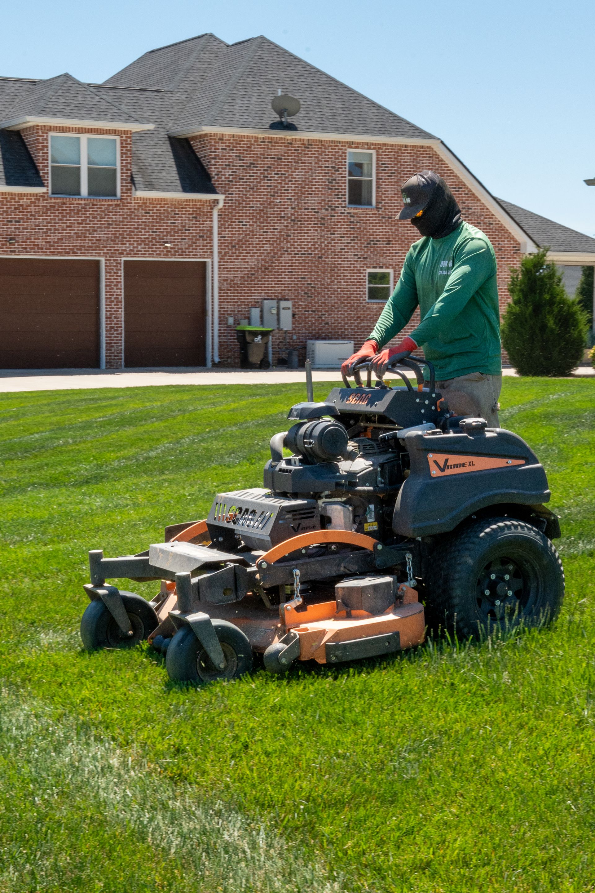 Person mowing a green lawn with a riding mower in front of a brick house