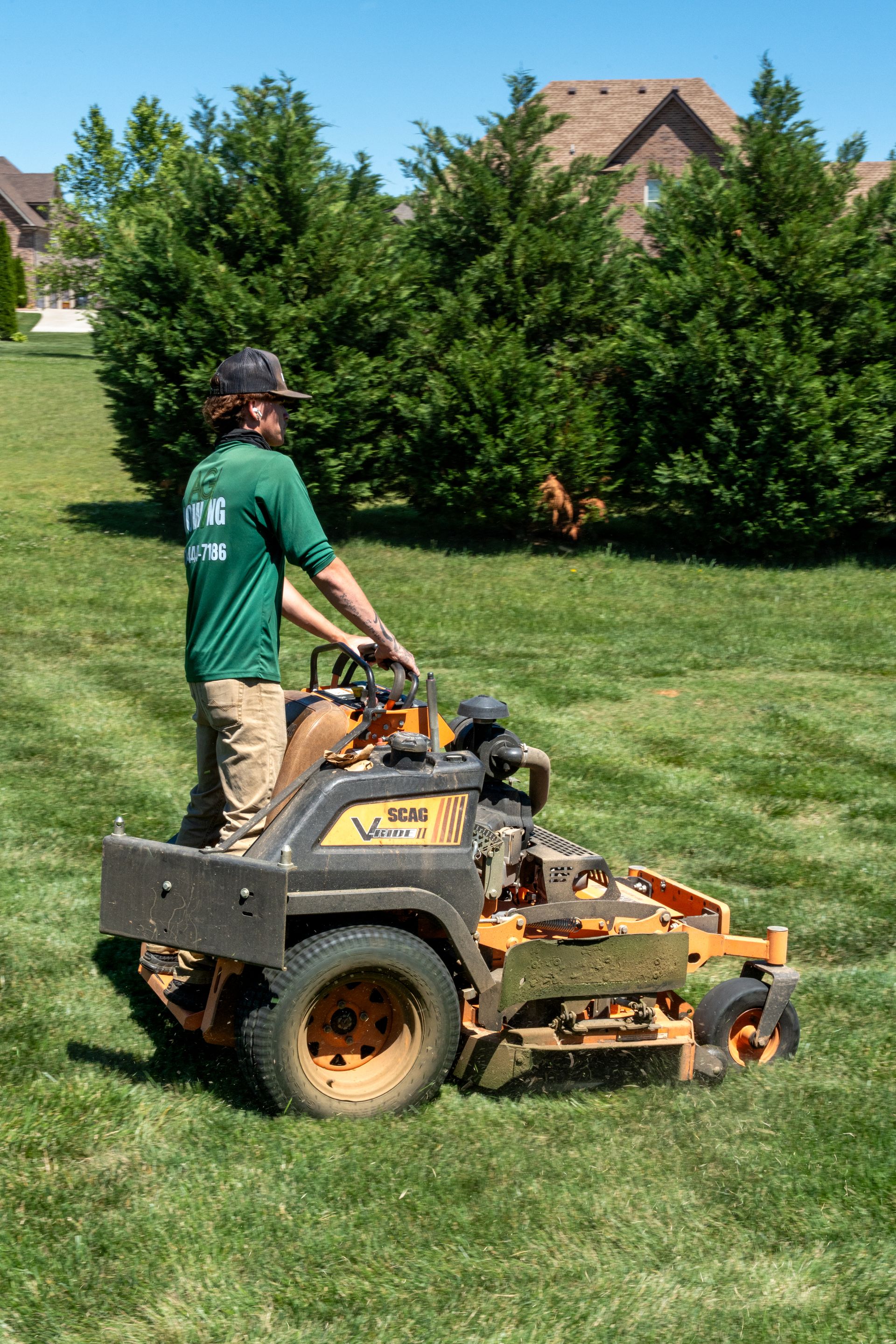 Person operating a yellow lawn mower on a grassy lawn near a hedge and houses