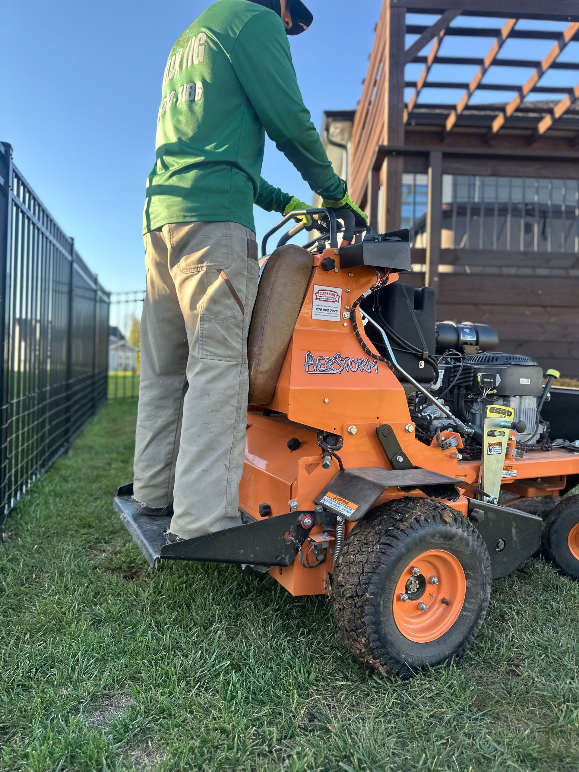 Man in green shirt operating an orange lawn aerator near a fence.