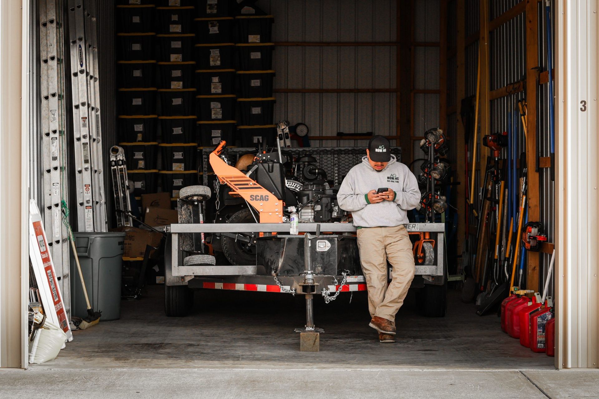 Man in gray sweatshirt and khaki pants leans on a trailer in a storage space, looking at his phone.
