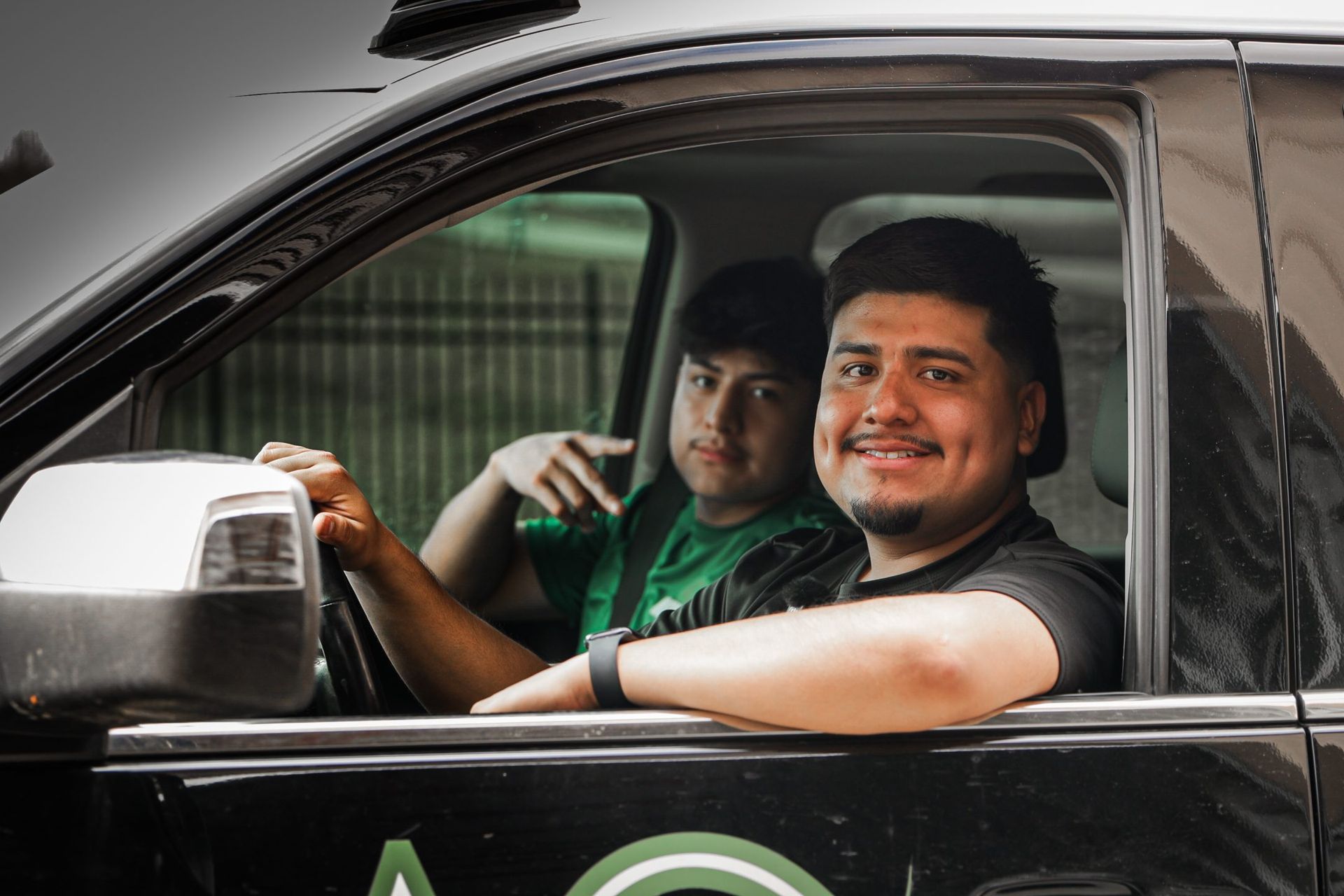 Two people in a black car; the driver smiles, the passenger gestures.