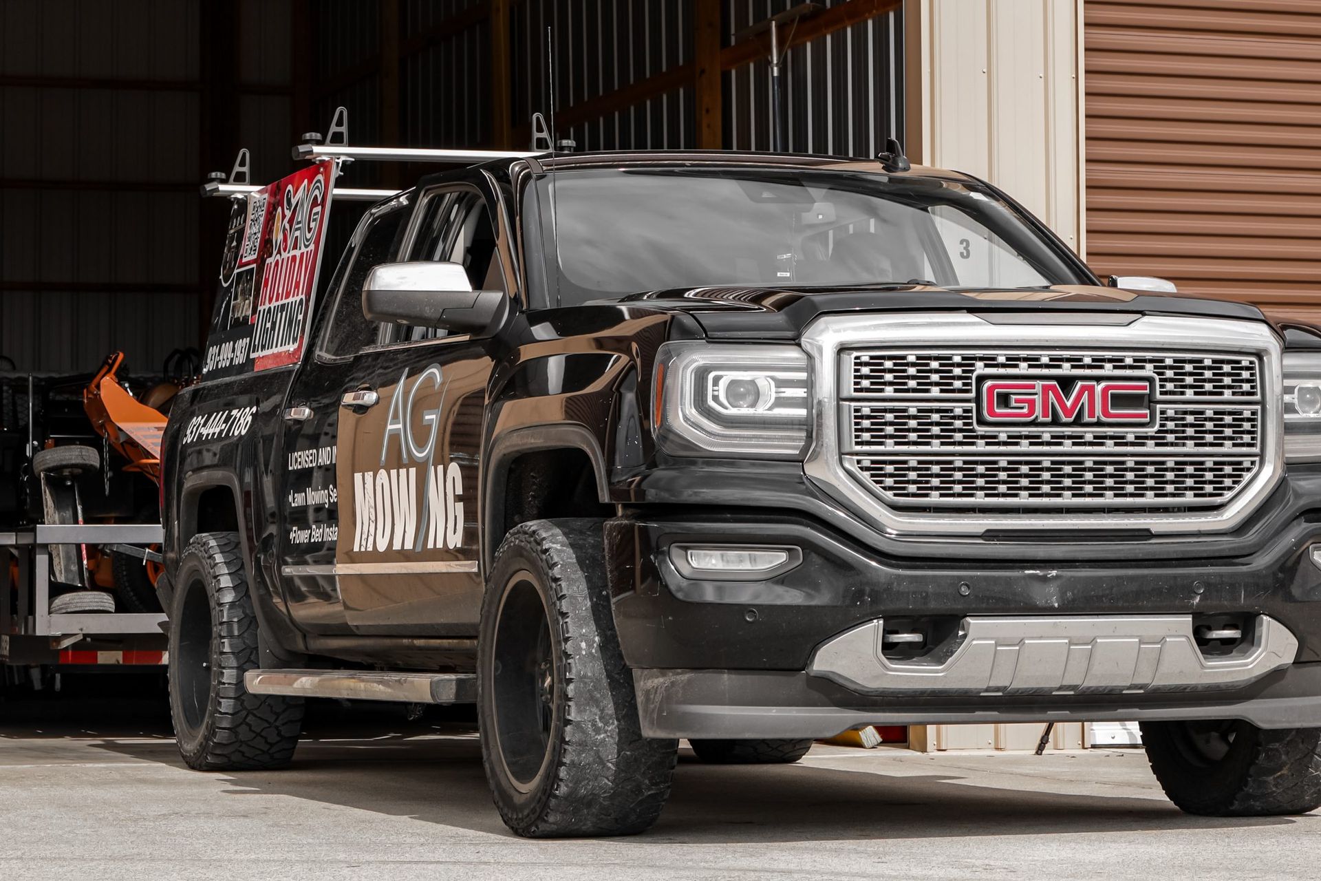 Black GMC truck with large tires parked in front of a building, pulling a trailer.
