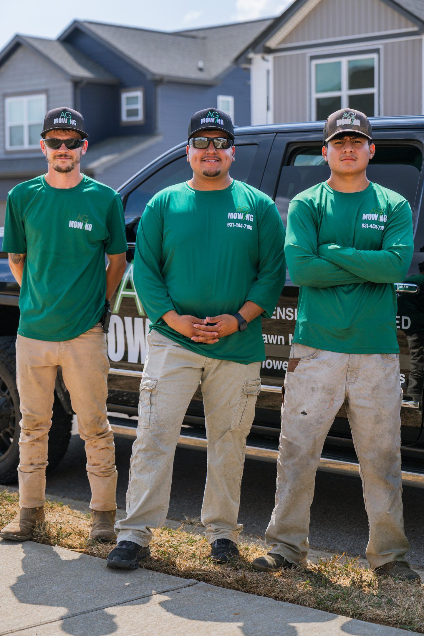 Three people in green shirts and tan pants stand in front of a black vehicle. They are in a suburban setting.