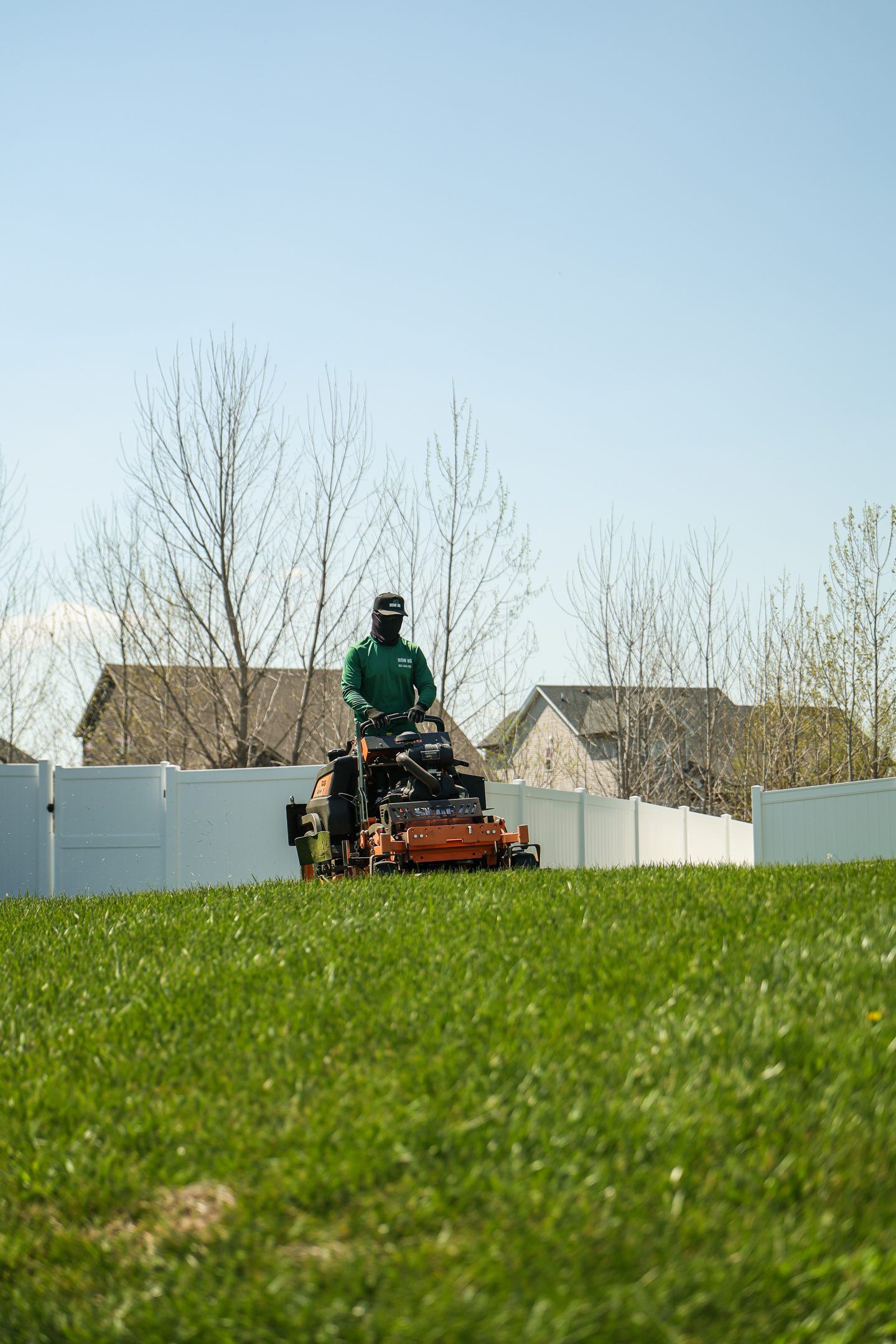 Person mowing a grassy lawn on a riding mower beside white fencing and houses under a clear sky