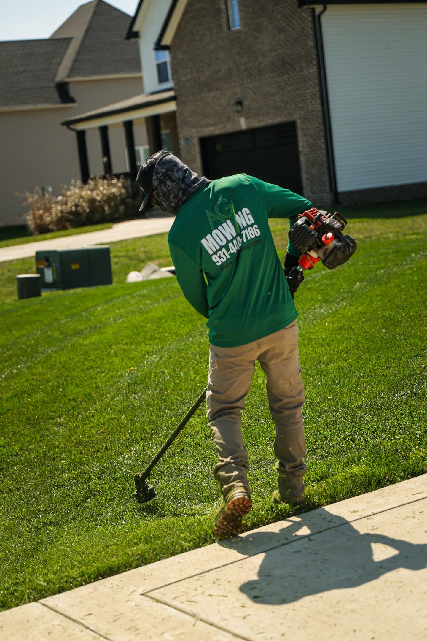 Person using a string trimmer on a grassy lawn beside a sidewalk in a suburban neighborhood.