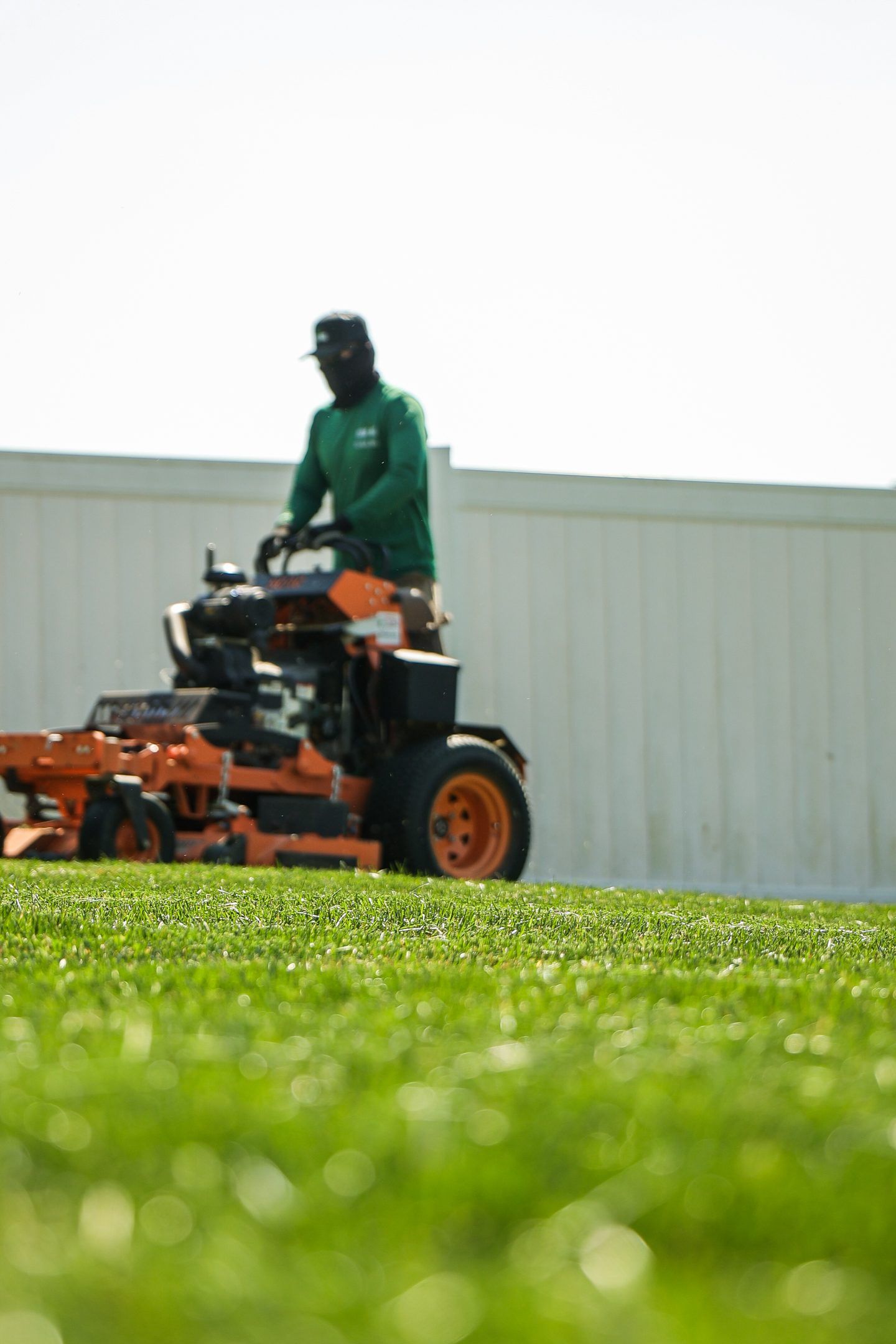 Person mowing grass with an orange riding lawn mower in a green field, with a white fence behind.