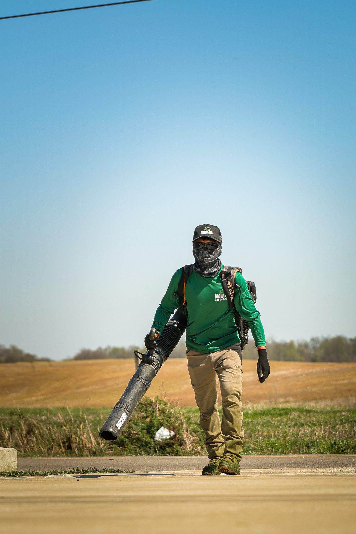Person in green shirt and tan pants walking outdoors with a leaf blower in a field under a clear sky