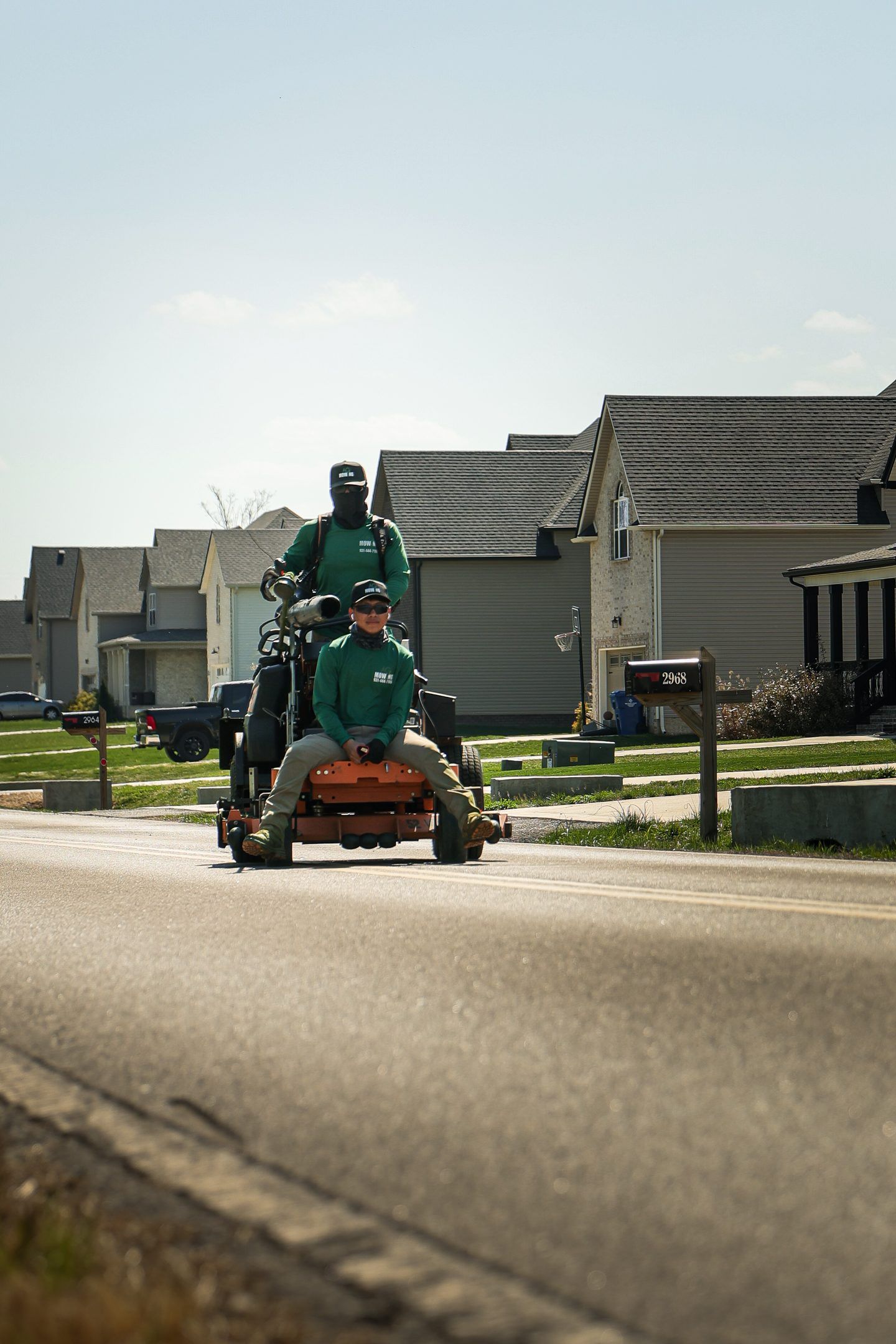 Two people on a lawnmower driving down a suburban street on a sunny day