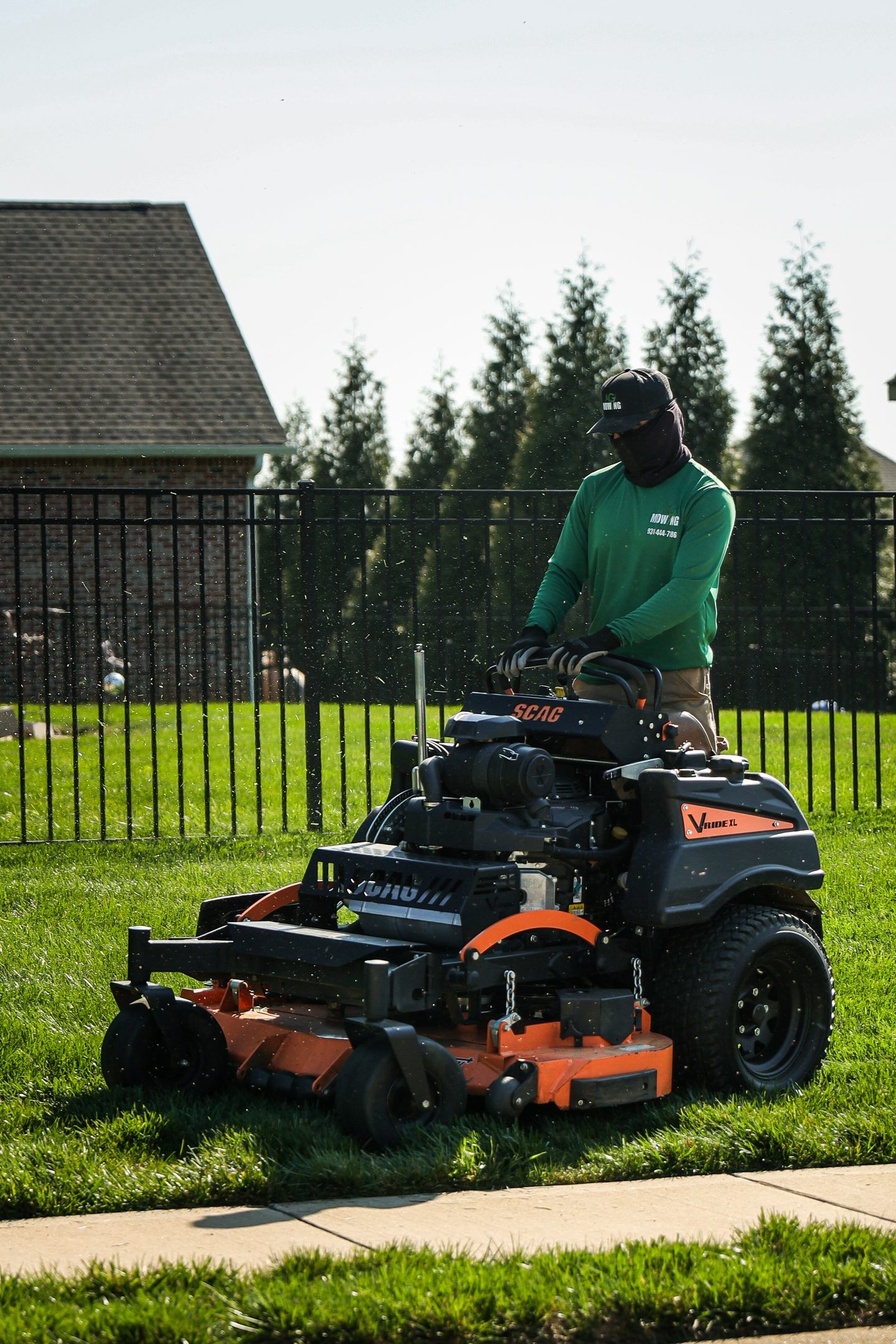 Person mowing grass with a riding lawn mower beside a fence and sidewalk.