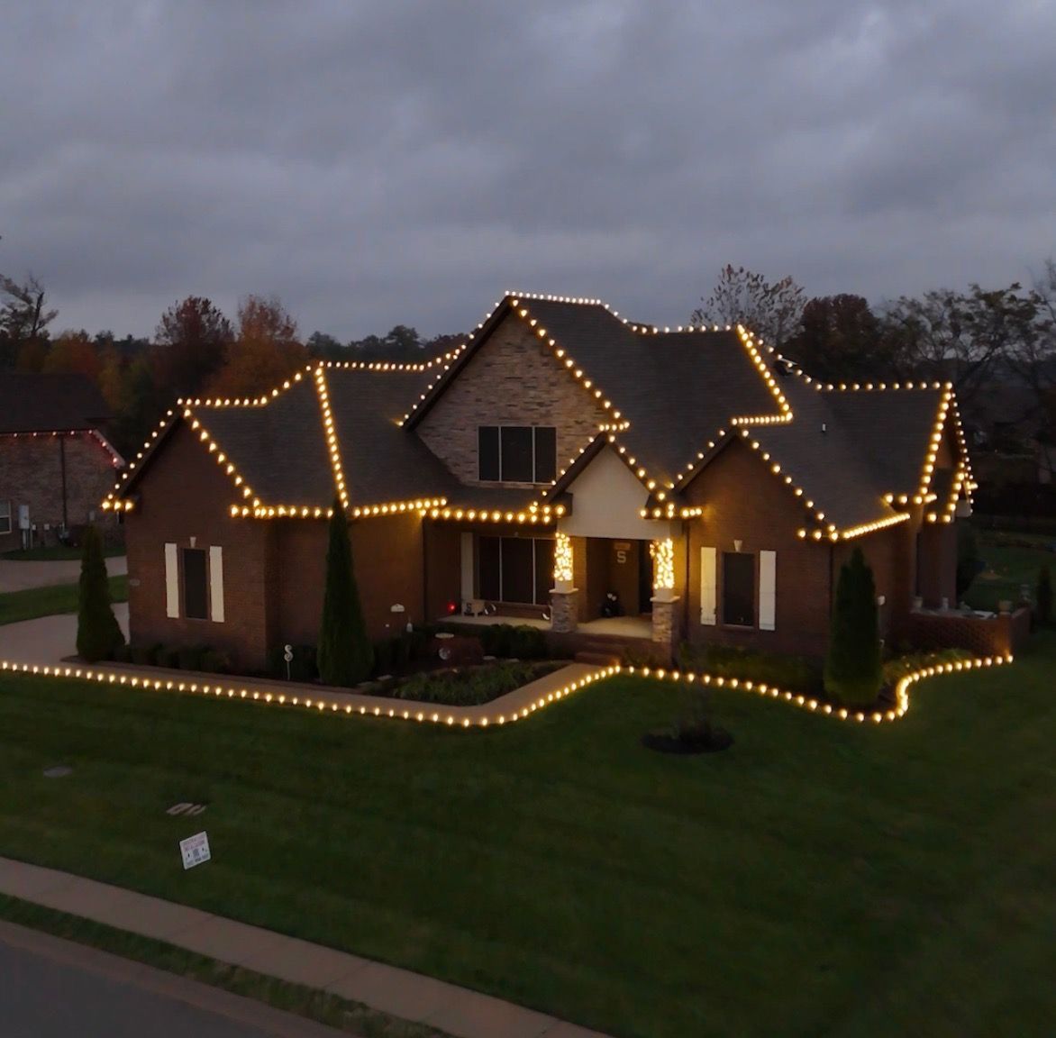 House decorated with yellow Christmas lights on roofline and around the yard, set against a cloudy dusk sky.