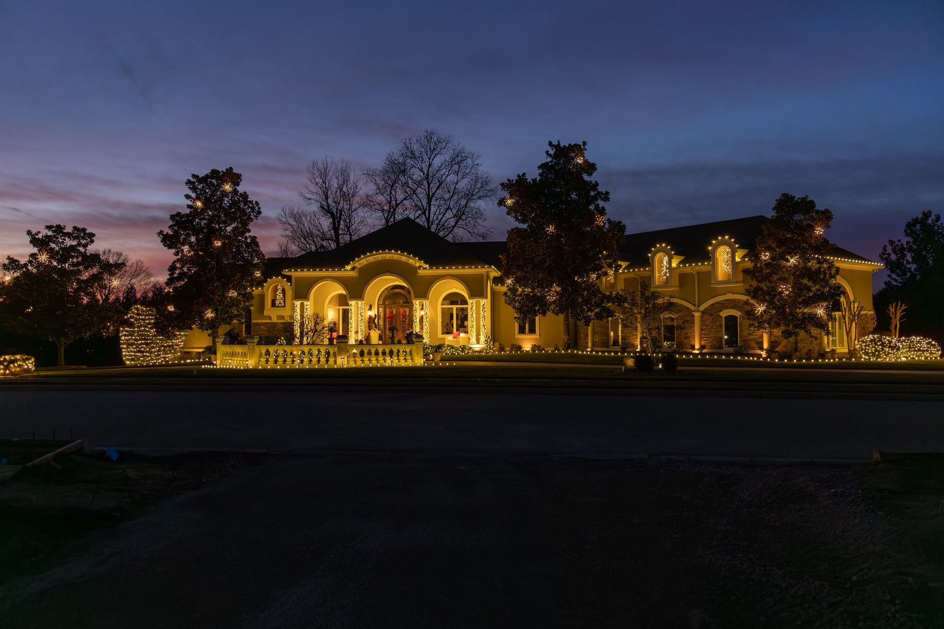 A large house with bright yellow lights at dusk. Trees and the sky are dark blue.