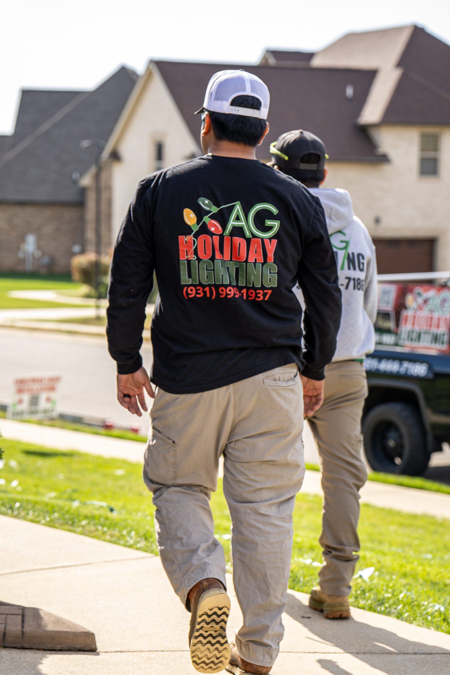 Two men walking on a sidewalk, wearing company logo shirts, near a suburban house.