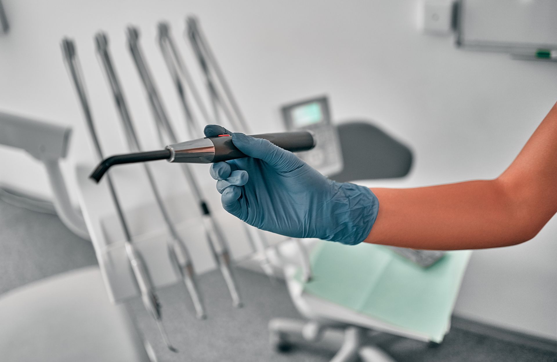 Gloved hand holding dental curing light in a dentist's office with dental tools in the background.