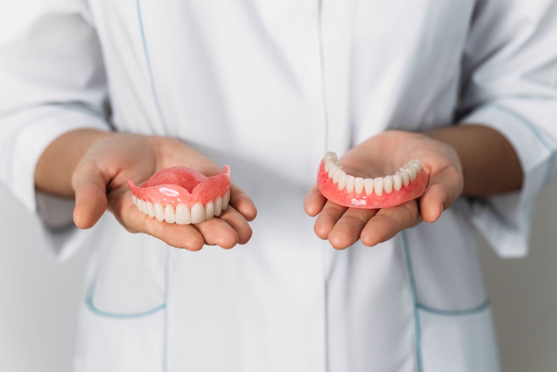 Person in white coat holding upper and lower dentures, pink with white teeth.
