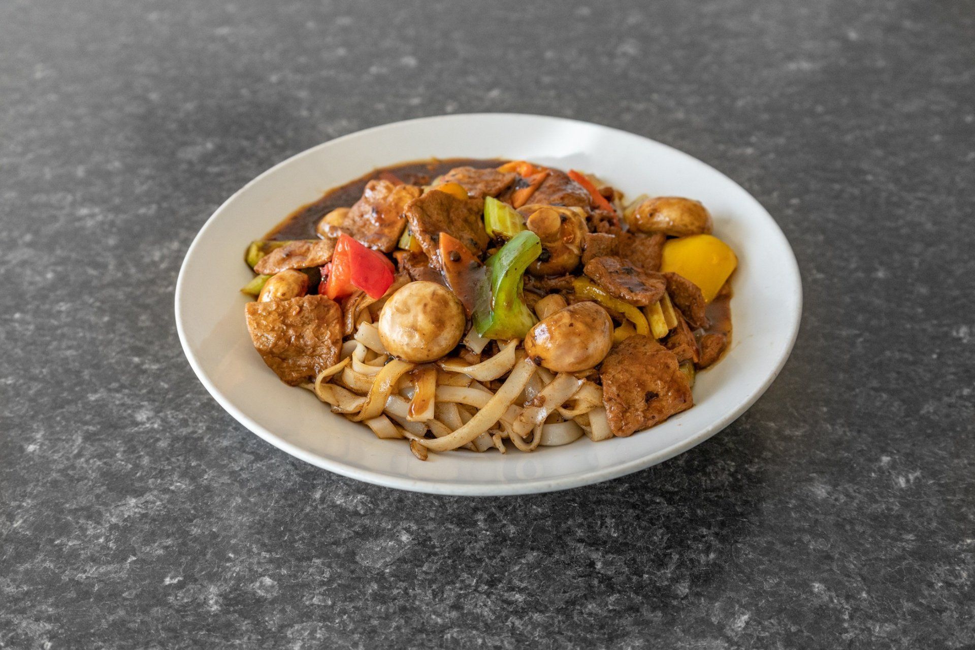Plate of noodles topped with a dark sauce, vegetables, and meat on a dark countertop.