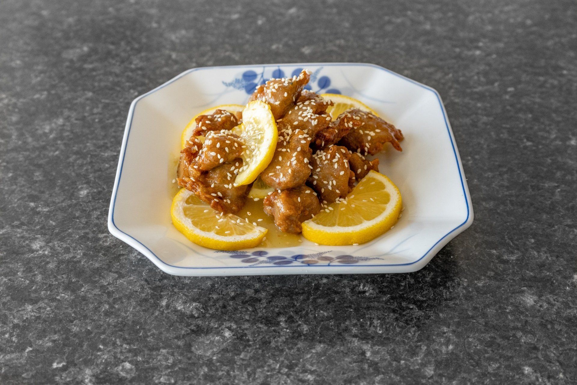 Sesame chicken with lemon slices on a white plate with blue floral trim, set on a gray countertop.