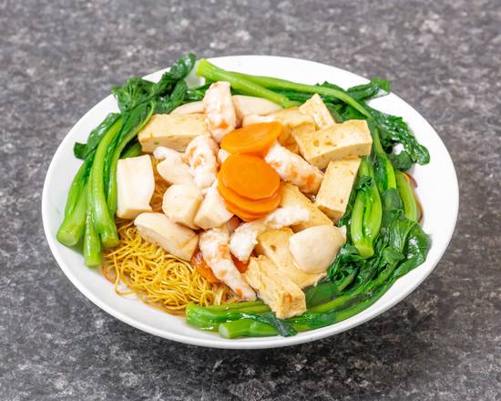 Noodles with vegetables, tofu, shrimp, carrots, and leafy greens in a white bowl.