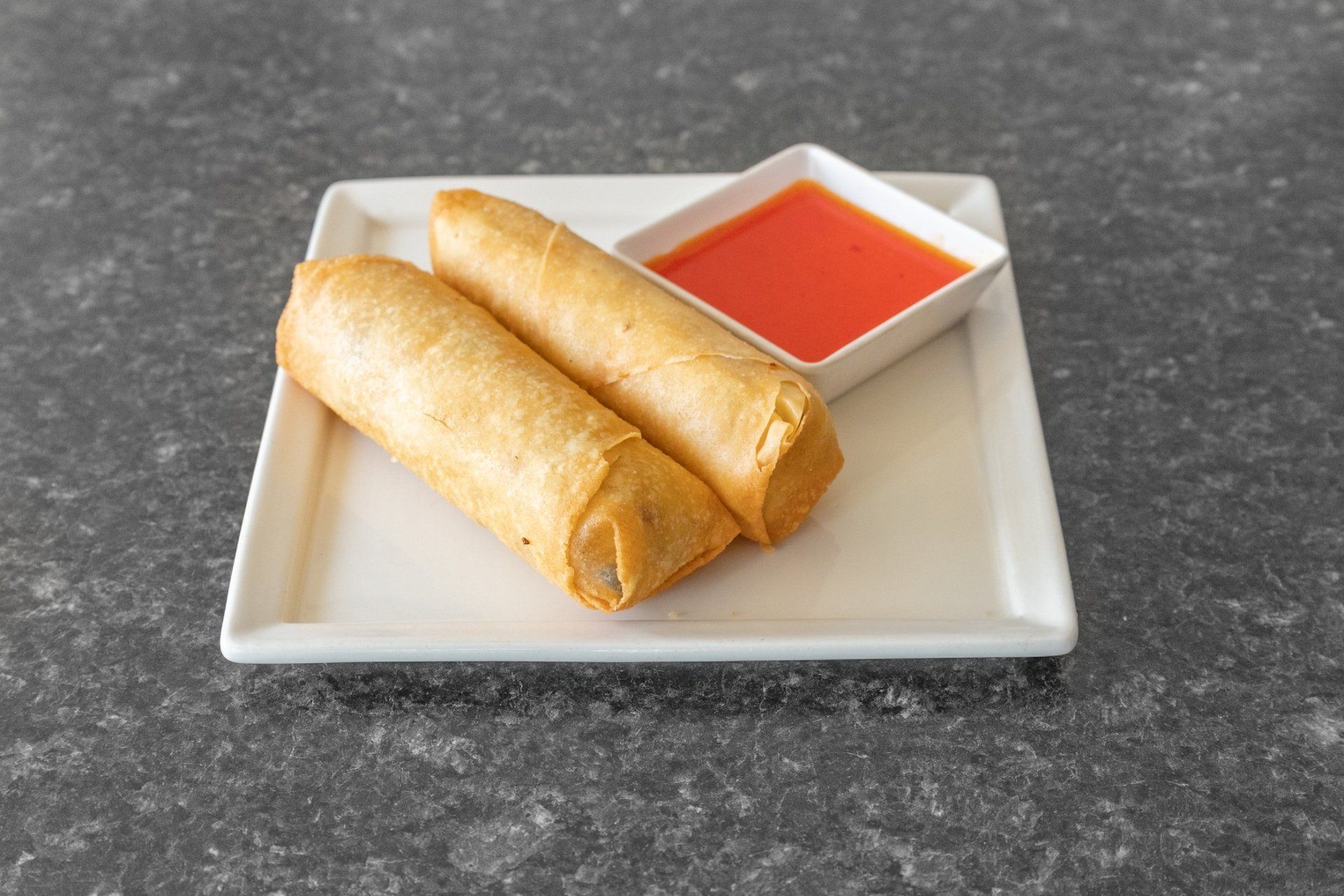 Two golden spring rolls with a side of sweet chili sauce on a white plate, gray surface.