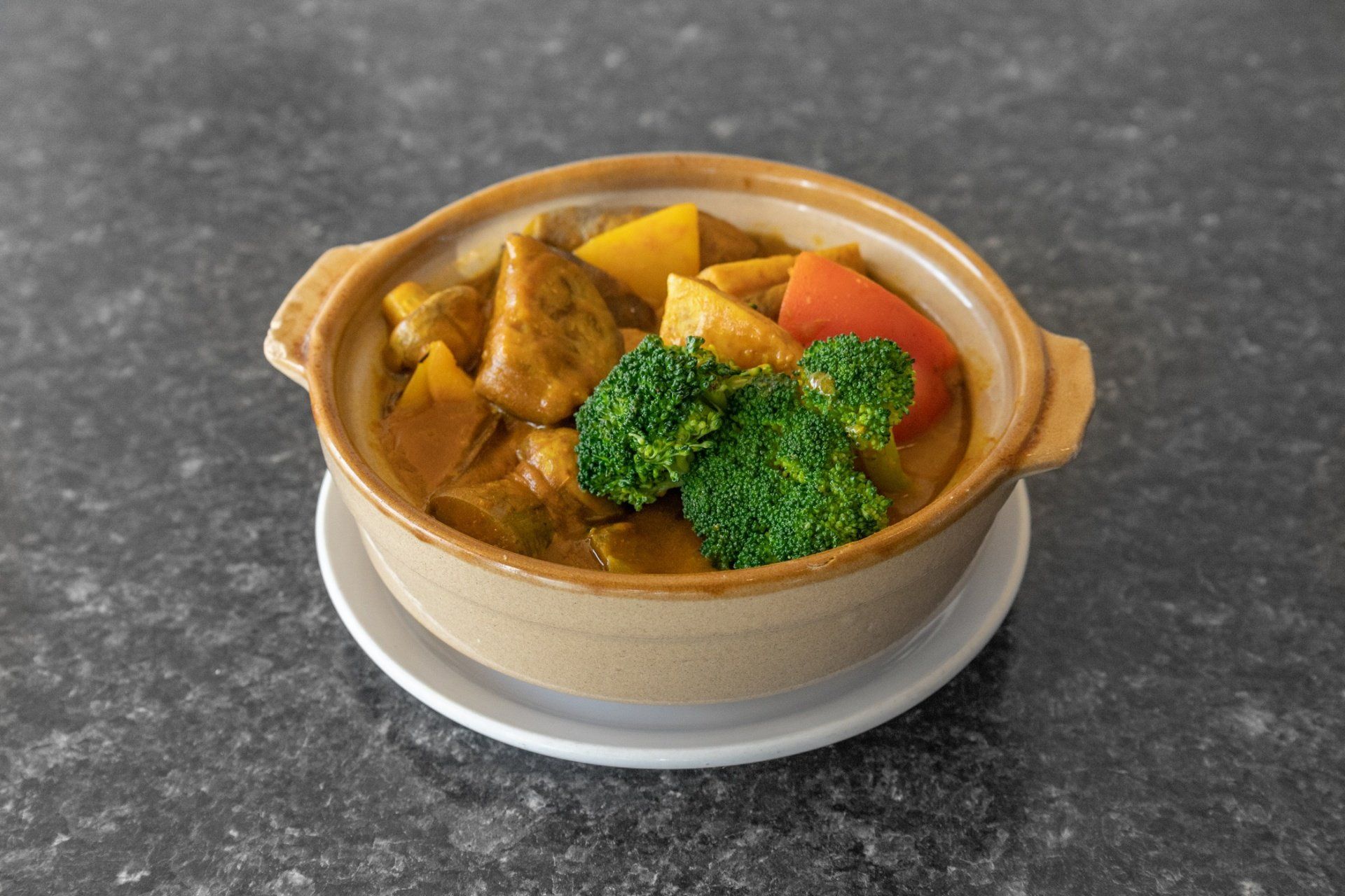 Curry dish in a ceramic bowl with broccoli, red pepper, and pumpkin on a white plate, dark gray countertop.