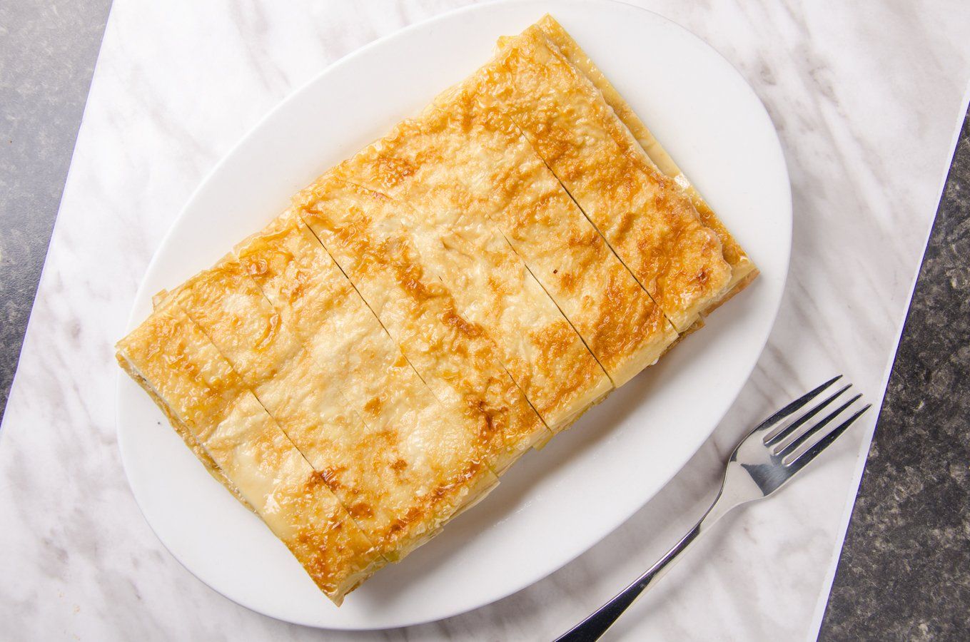 Golden-brown, square pastry on a white plate, cut into squares. Silver fork on the side.