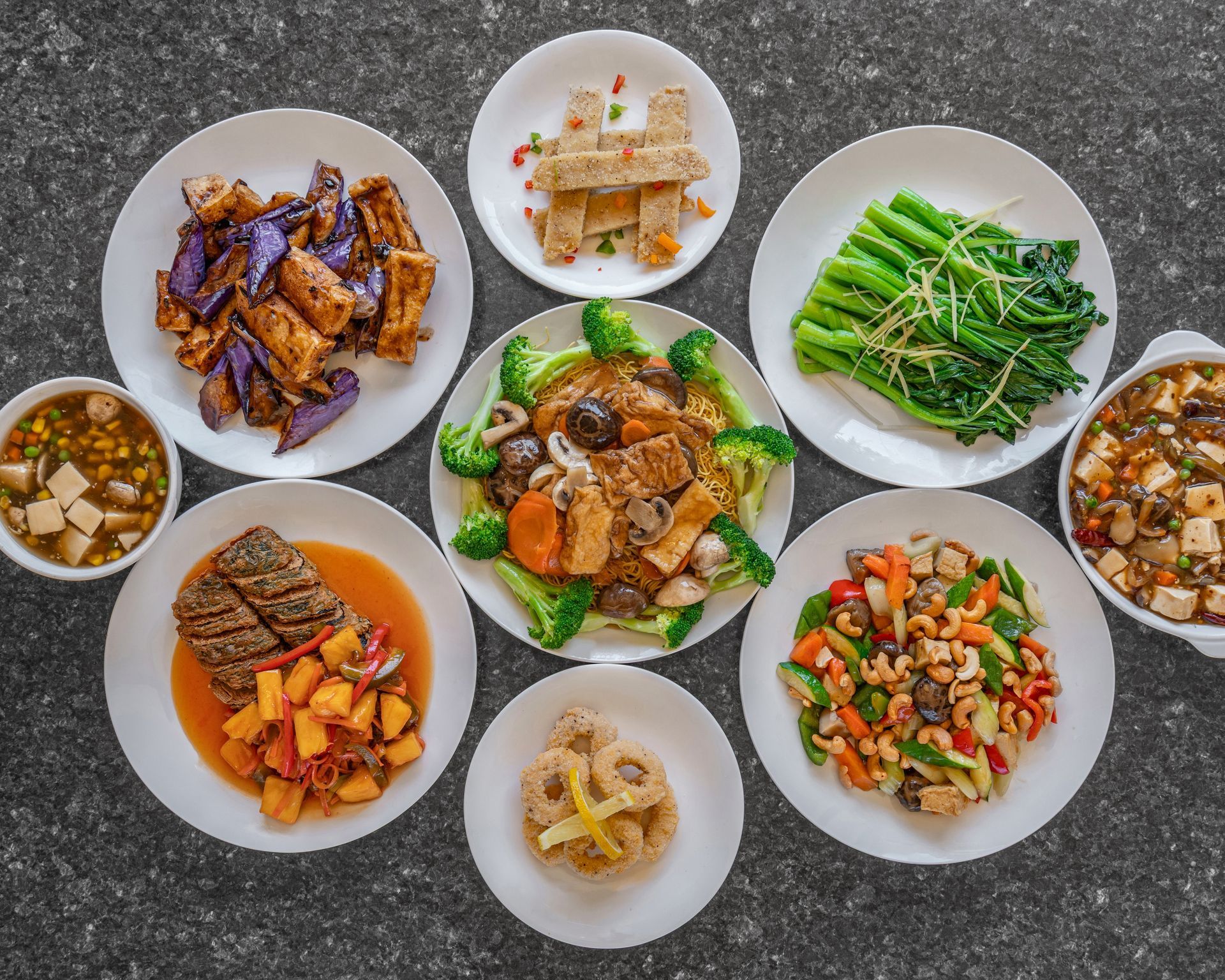 Variety of Chinese dishes on plates, including tofu, vegetables, and meat, arranged on a gray surface.