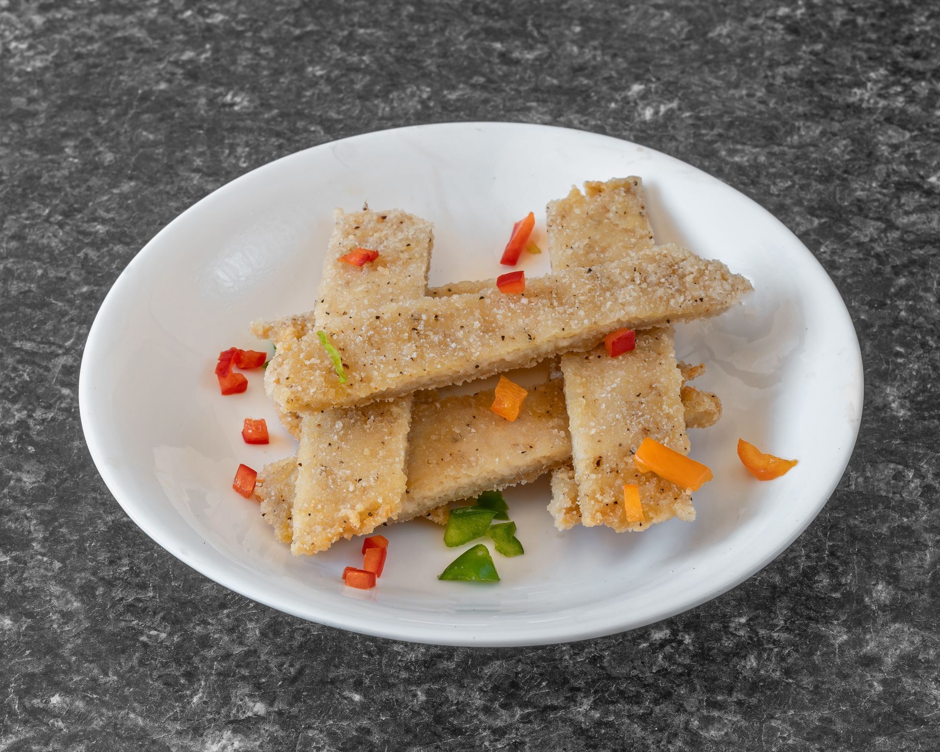 Breaded sticks on a white plate with diced red, green, and orange peppers; gray countertop in the background.