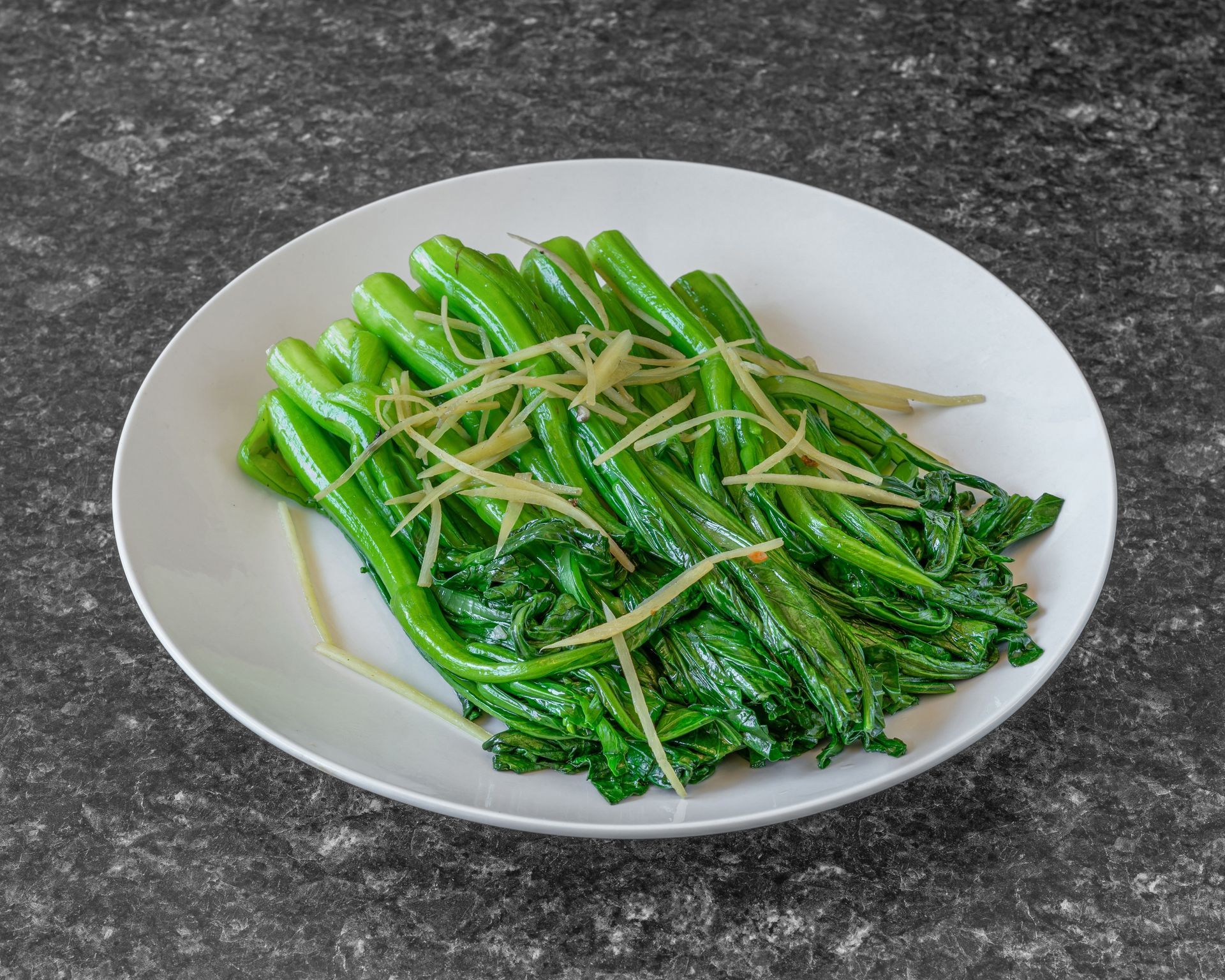 Stir-fried Chinese broccoli topped with ginger slivers on a white plate, set on a dark surface.