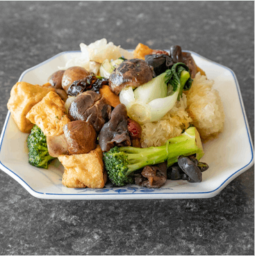 Vegetarian dish on a white plate with tofu, broccoli, mushrooms, bok choy, and black fungus against a gray backdrop.