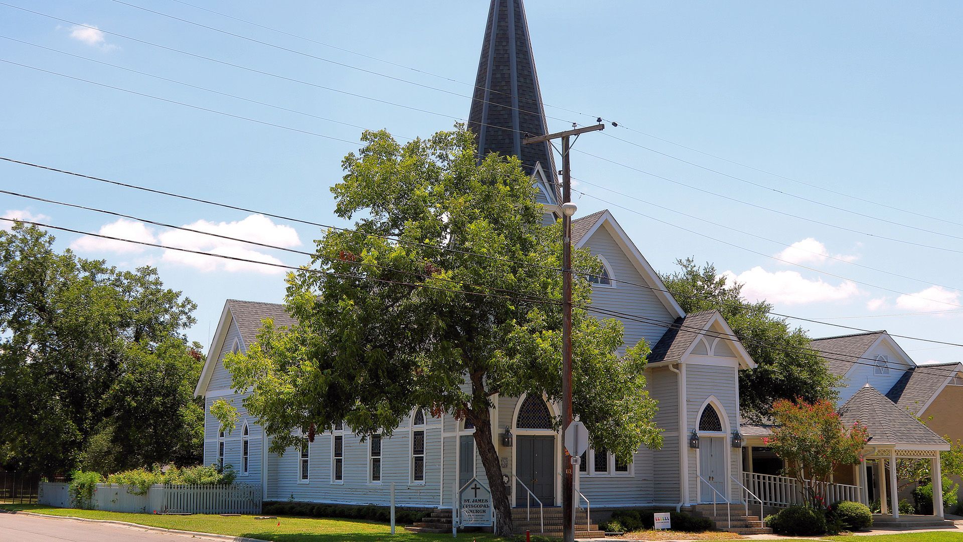 White church with a tall black steeple and trees in front under a blue sky.