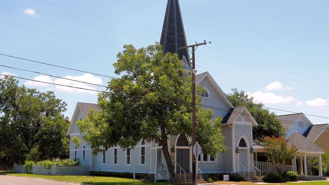 White church with tall dark steeple and stained-glass windows under a blue sky, partly shaded by a tree