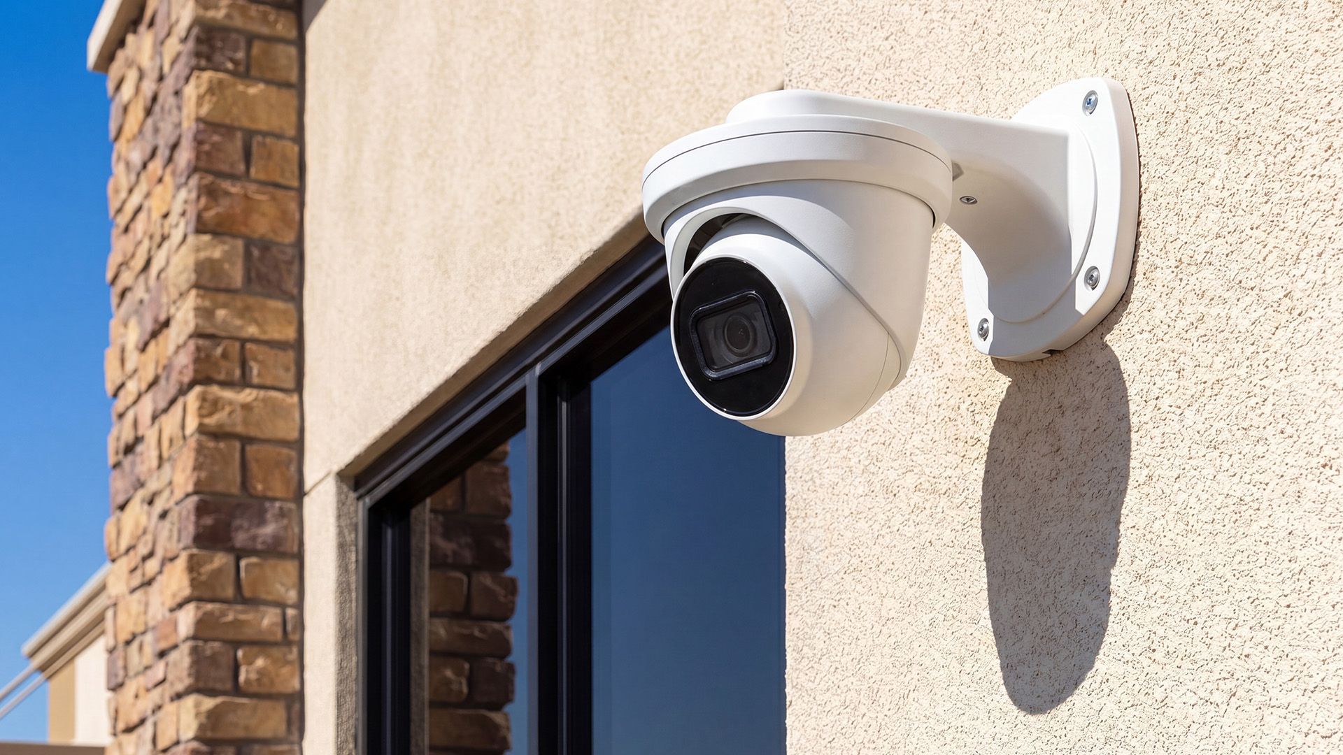 White security camera mounted on a beige house exterior beside a window and brick wall