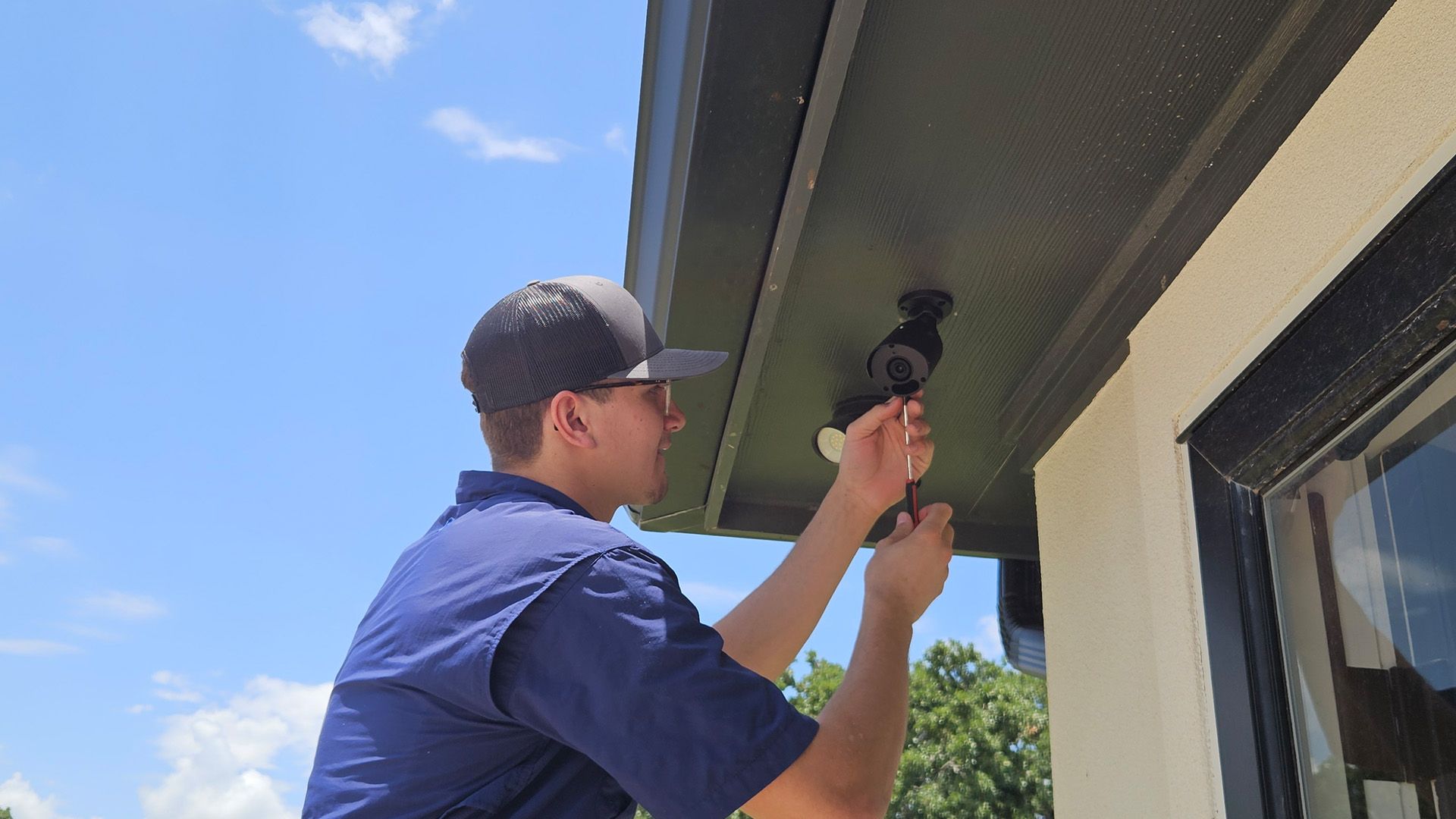 Person in a blue shirt and cap using a drill near a house roofline outdoors.