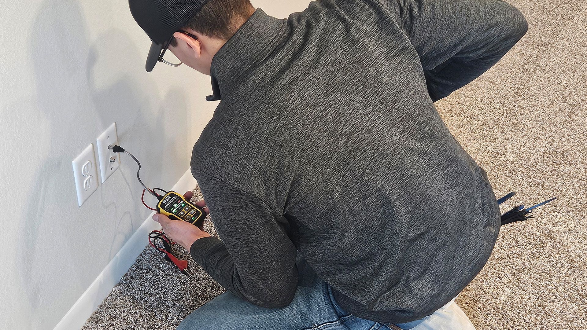 Person kneeling by a wall outlet, plugging in wires on a speckled floor.