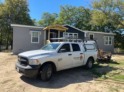 White work truck parked in front of a small gray house with a covered porch and trees behind it