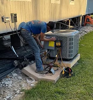 Technician working on an outdoor HVAC unit beside a house, with tools and cables on the ground