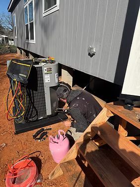 Worker kneeling beside HVAC unit under a raised gray house, with tools and buckets on reddish dirt.
