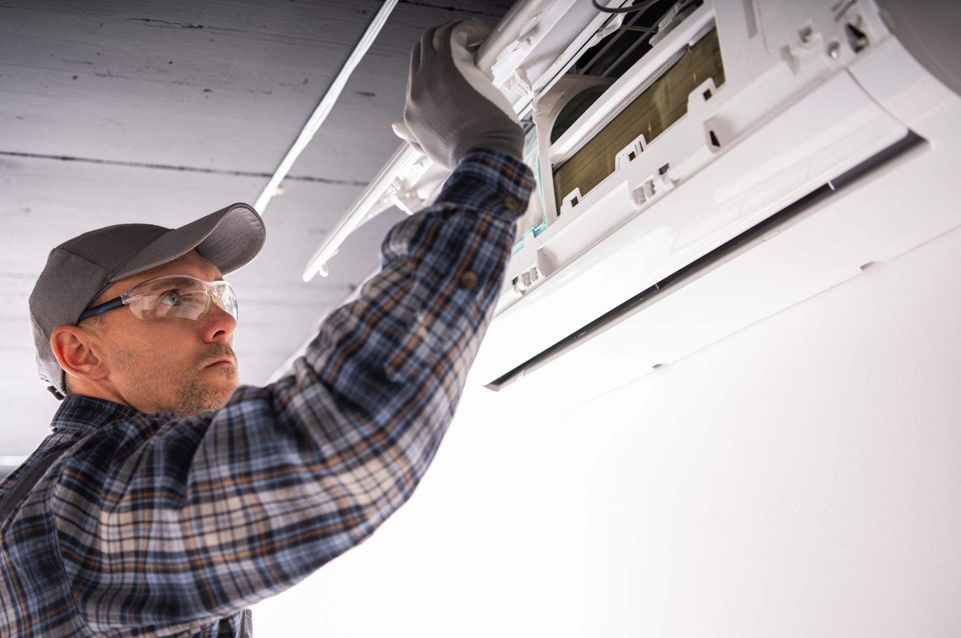 Worker in plaid shirt and cap installing a ceiling light fixture with a screwdriver.