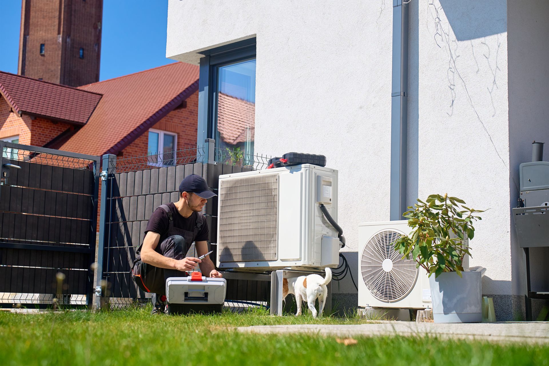 Person kneeling beside outdoor HVAC units in a backyard, working with tools near a small dog