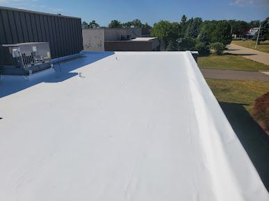 White flat commercial roof on a sunny day with a HVAC unit and trees in background.