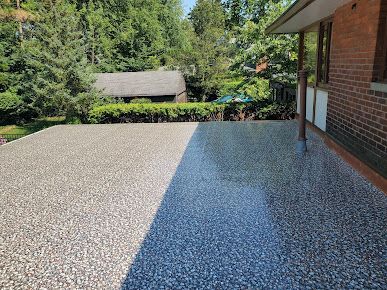 Concrete patio with pebble finish; partial shadow, brick wall, and trees in the background.