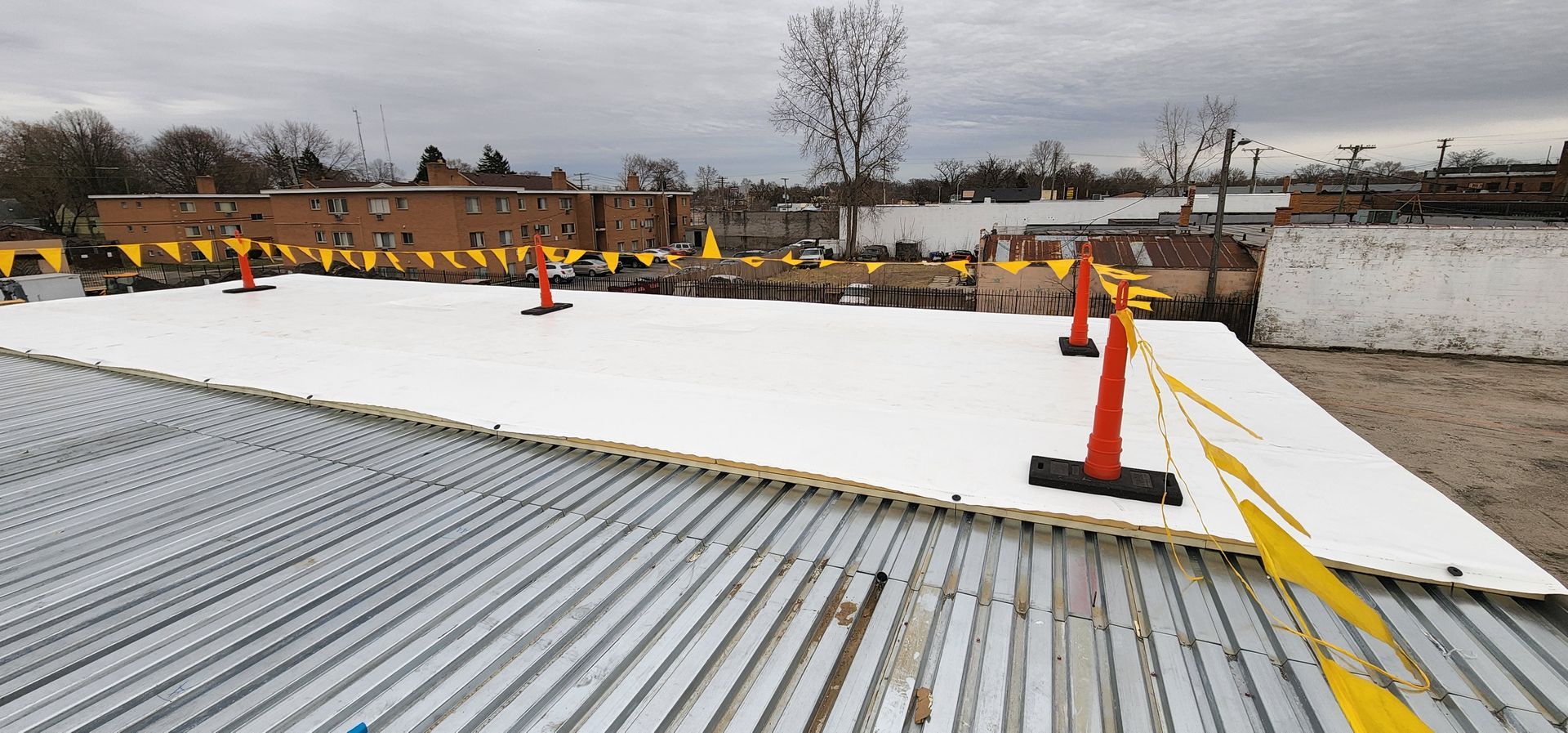 A roof with white insulation panels and safety cones, yellow caution tape, a brick building in the background.