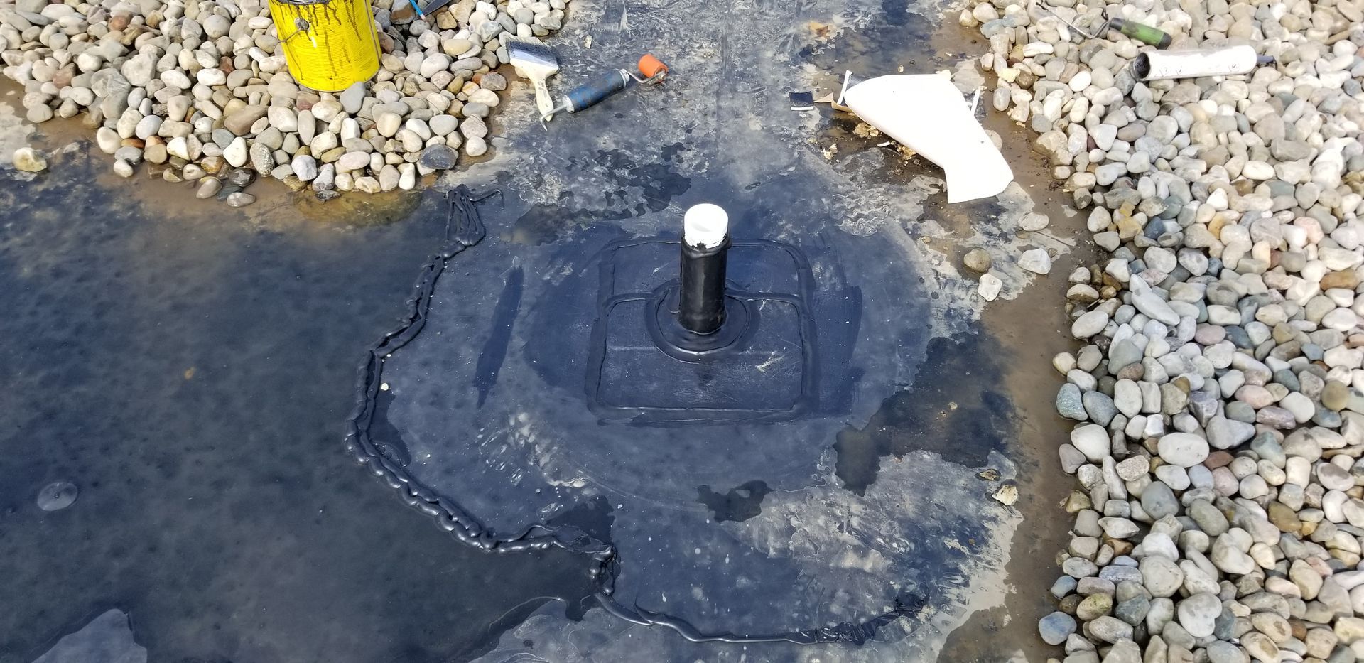 A black pipe sticking out of a dark tar-like substance. The surface is surrounded by water and gravel.