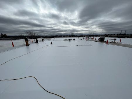 Workers installing white roofing membrane on a flat roof under a cloudy sky, with orange cones.