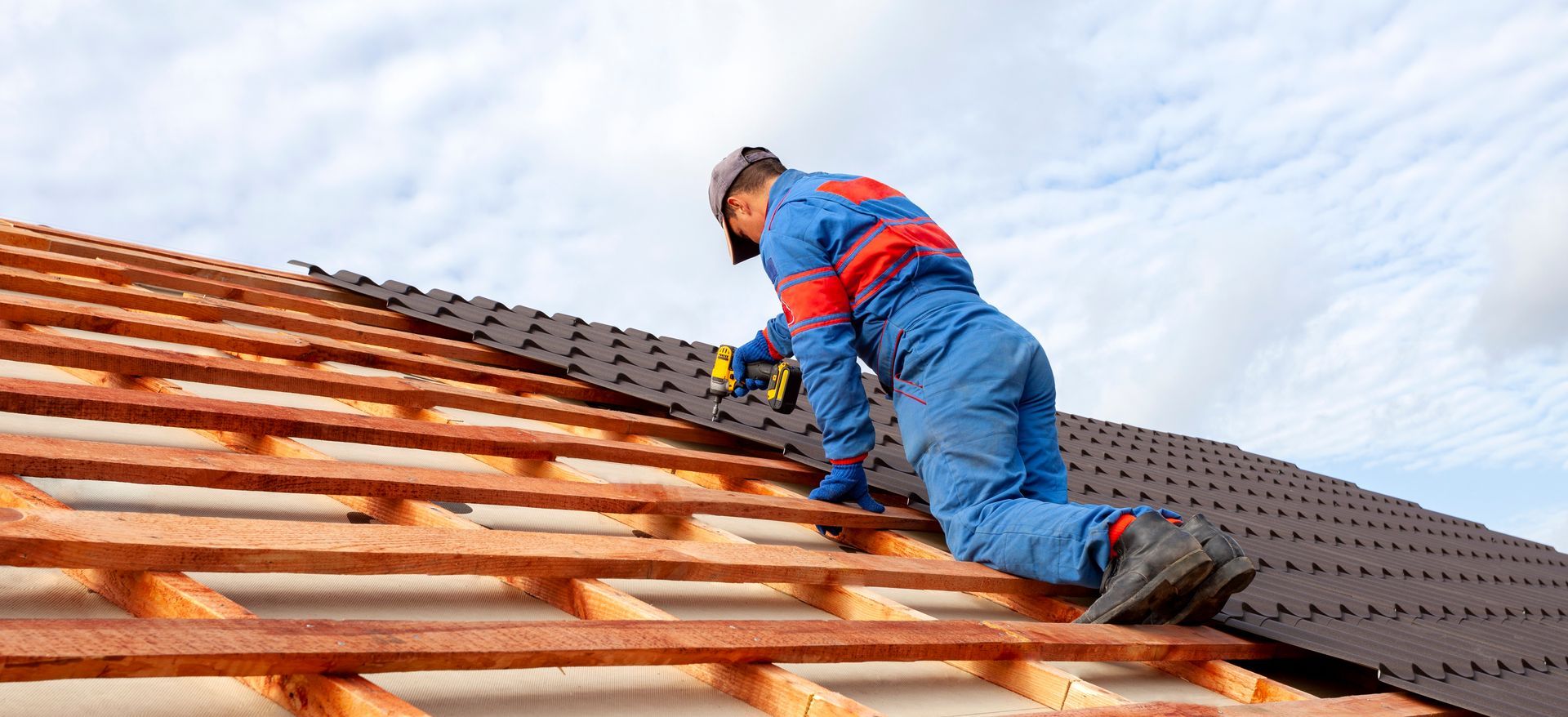A construction worker in blue coveralls installing shingles on a roof.