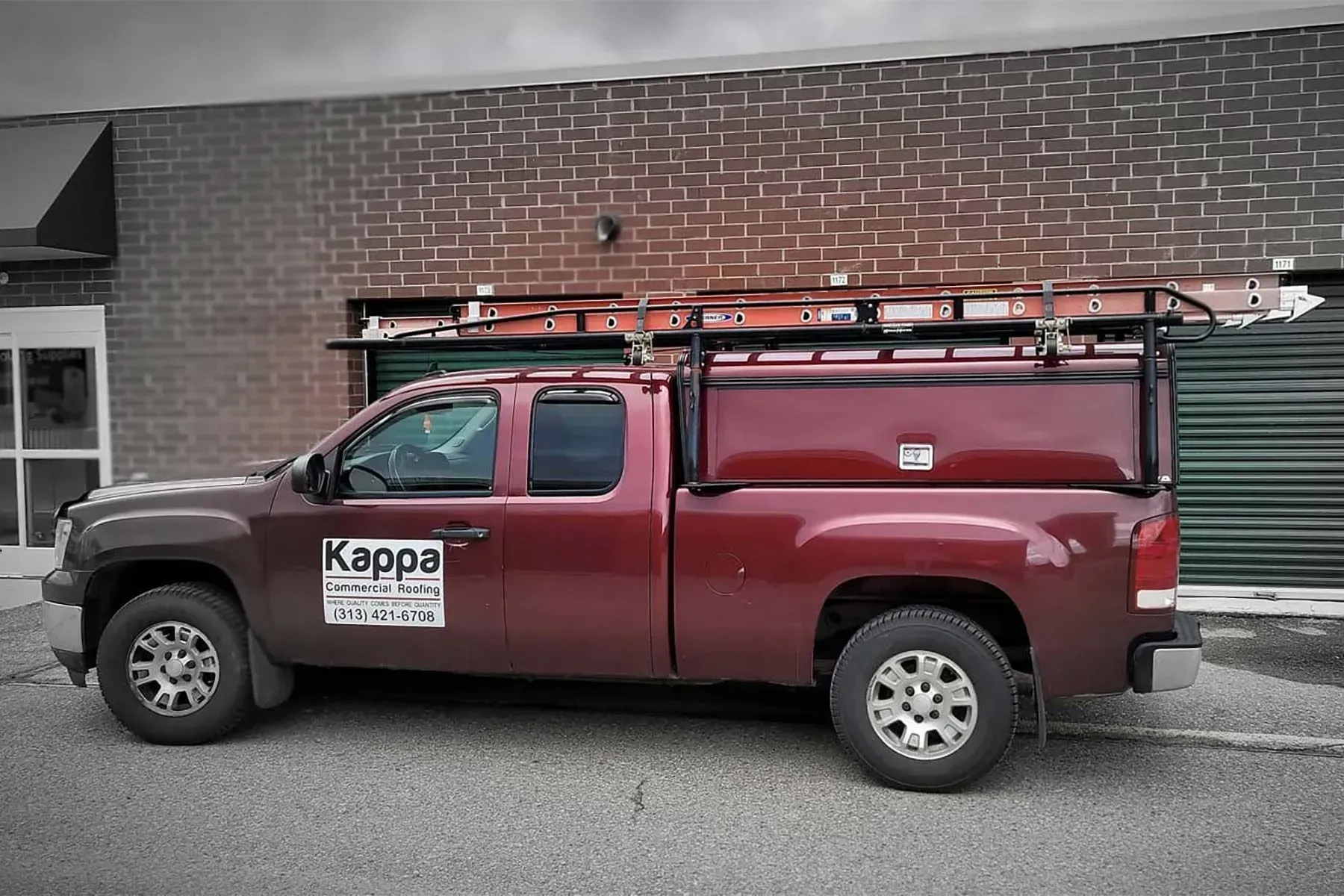 Red pickup truck with ladder rack, storage box, parked in front of a brick building.