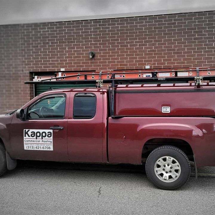 Red truck with roof rack and ladder, parked in front of a brick building; 