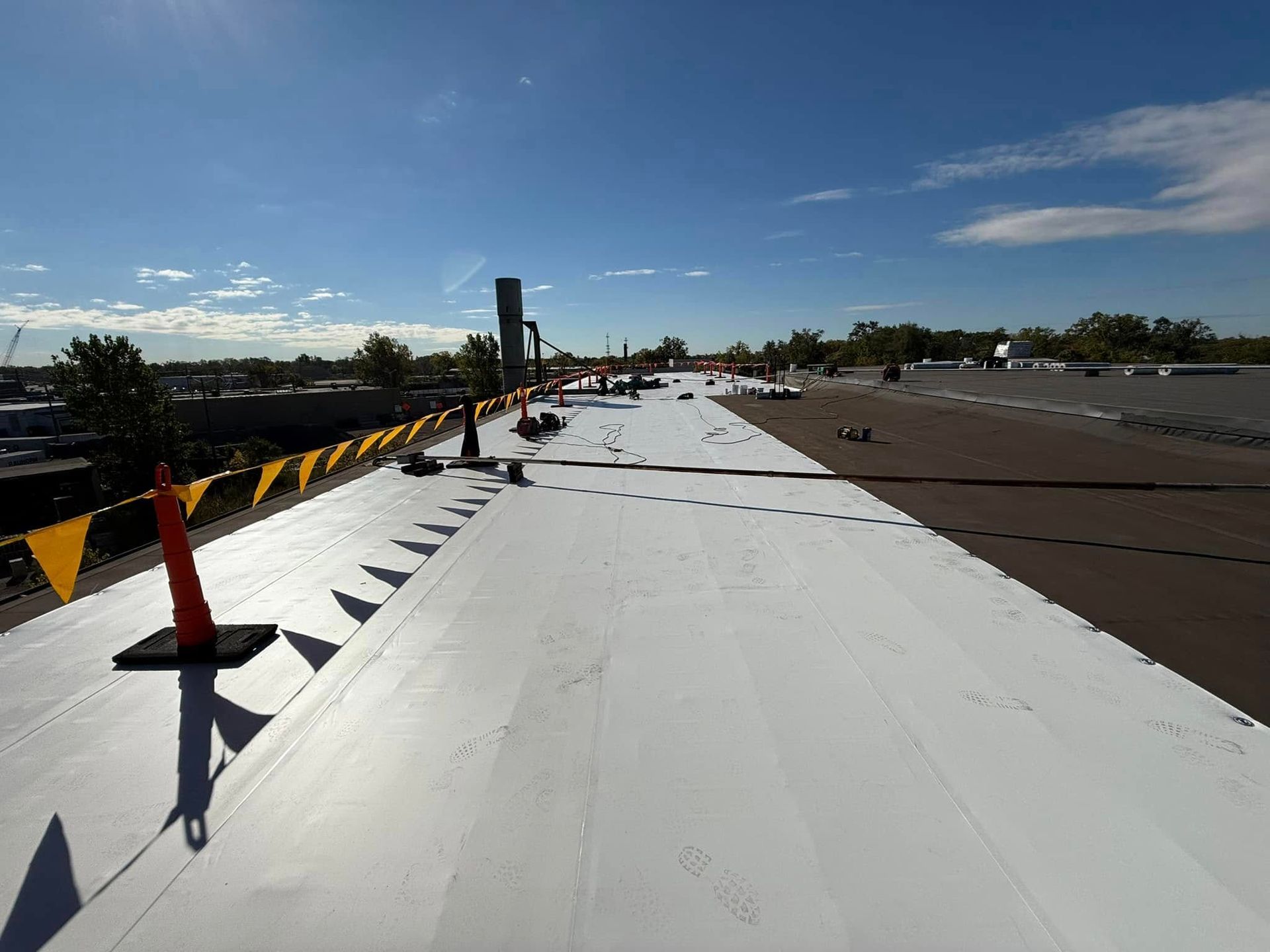 White commercial roof under construction with orange cones and yellow flags. Clear blue sky.