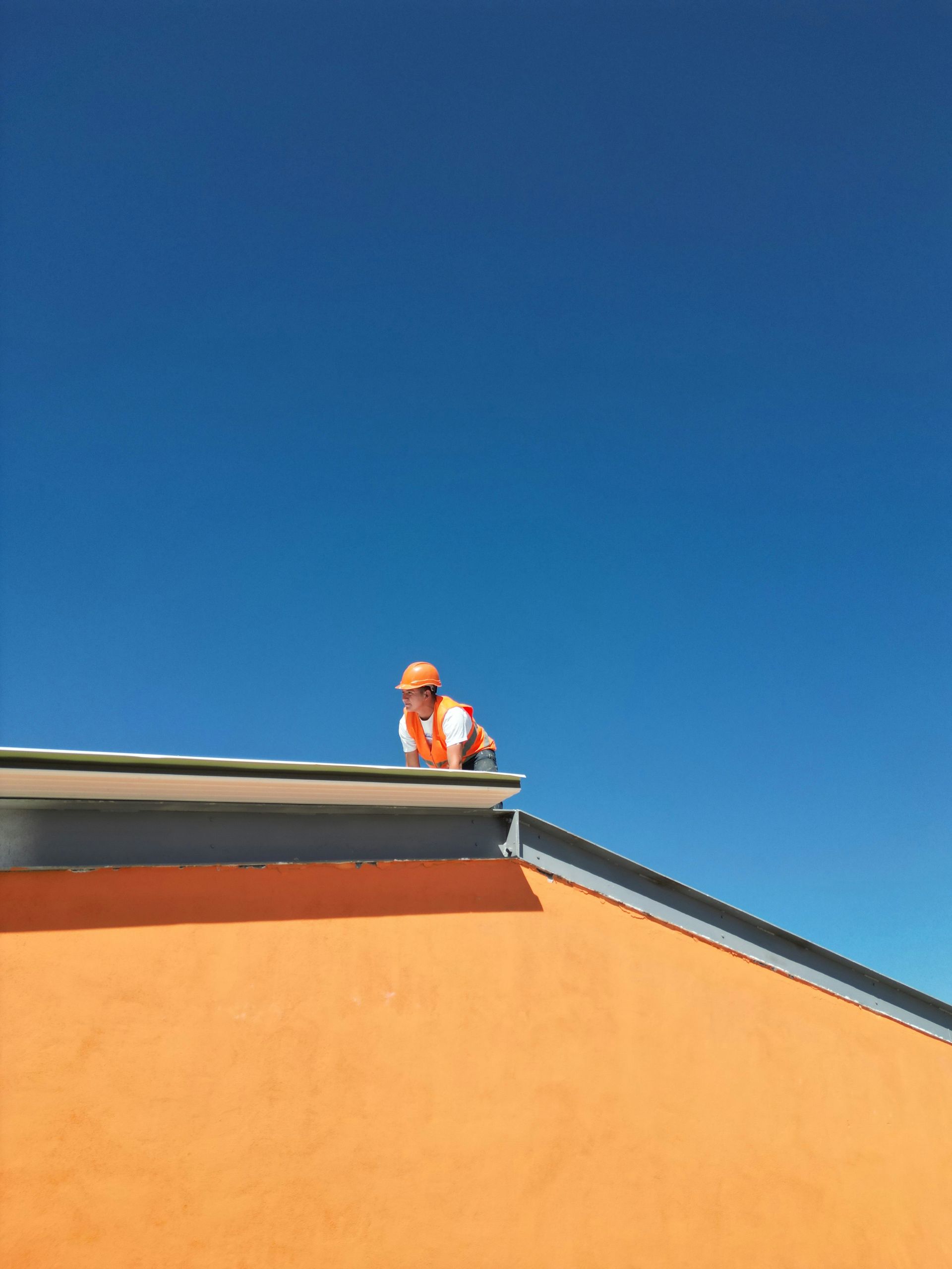 Construction worker on orange building roof against a clear blue sky.