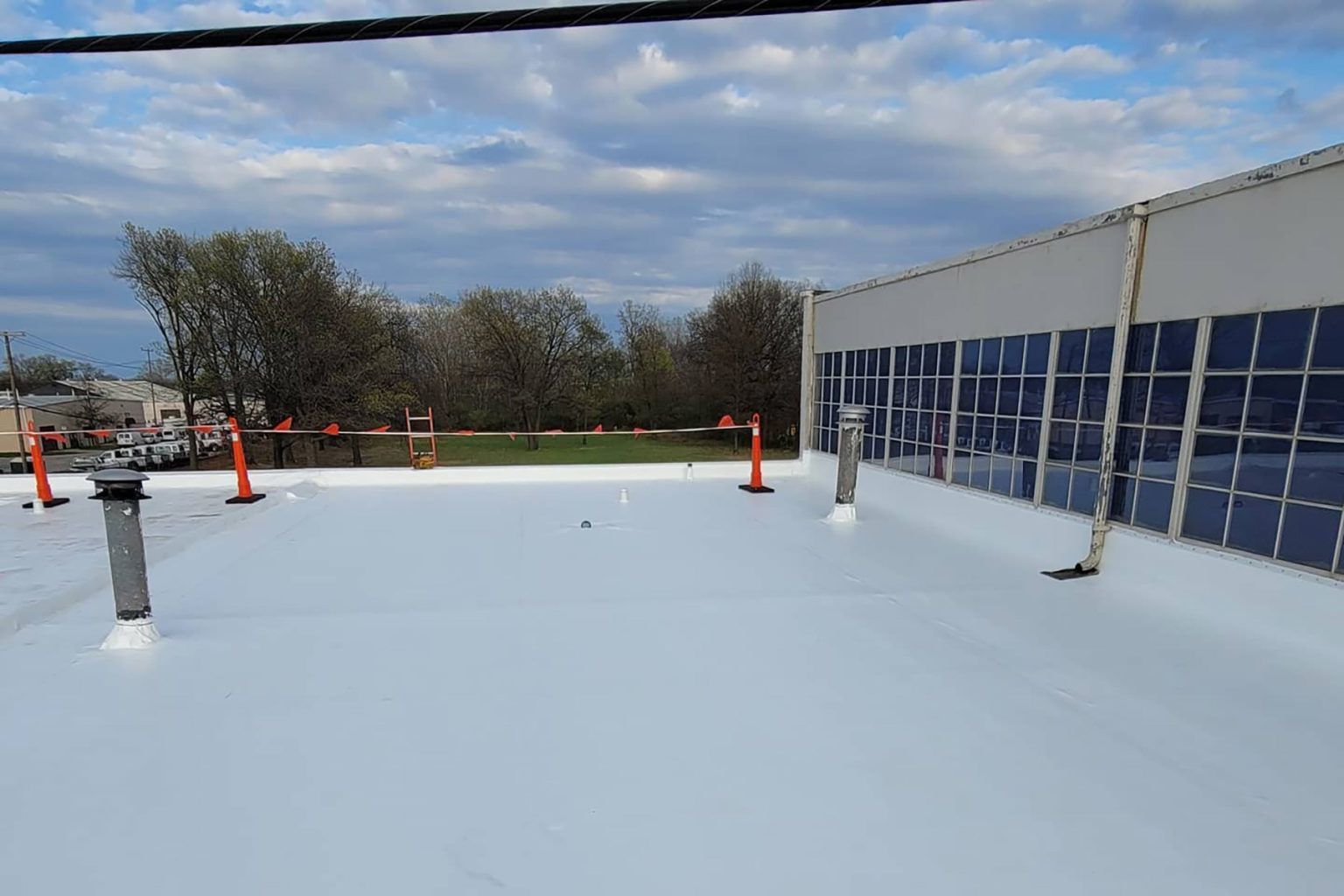 White commercial roof with vents and cones; building on the right, trees in the background under a cloudy sky.